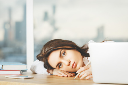 Portrait of beautiful young woman laying head on wooden office desk with blurry city view in the background. Overworked conceptの写真素材