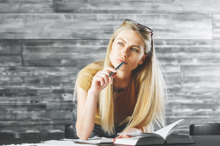 Pretty thoughtful young woman reading book and doing paperwork in office interior with wooden desk and background. Research conceptの写真素材