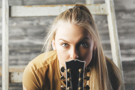 Portrait of pretty young woman with guitar neck sitting on wooden stairs. Blurry background. Music, hobby, leisure conceptの写真素材