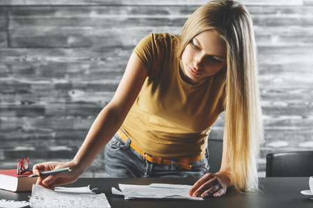 Young woman doing paperwork at office desk with book, coffee cup and other items. Blurry wooden wall in the background. Project, contract, document conceptの写真素材