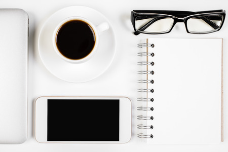 Above view and close up of white office desk with empty smartphone, spiral notepad, coffee cup, laptop and glasses. Mock up, Workplace conceptの写真素材