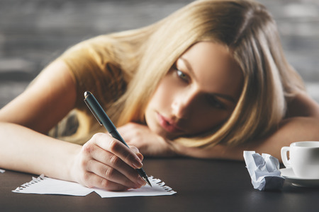 Close up portrait of tired young lady writing on paper sheets placed on desktop with coffee cup. Blurry wooden wall backgroundの写真素材