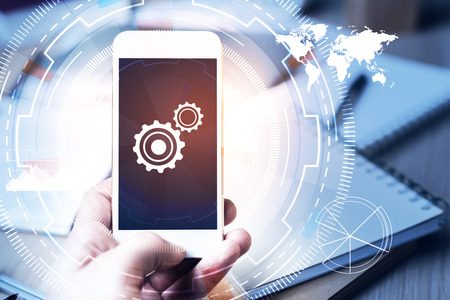 Hand holding smartphone with cogs on blurry workplace with tech pattern background. Settings concept. Double exposureの写真素材