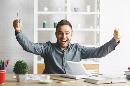 Happy young european businessman celebrating success at modern office deskの写真素材