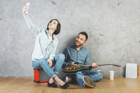 Attractive man and woman sitting on wooden floor with guitar, coffee cup, shopping bags and other items. Concrete wall background. Lifestyle, hipster, young, modern conceptの写真素材