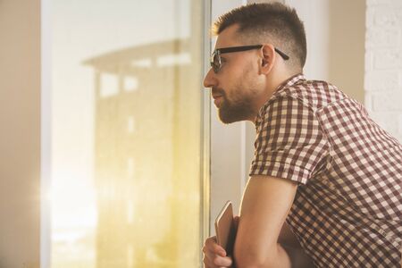 Side view of young businessman with smartphone looking out of window with city view, bright sunlight and copy space. Research and communication concept の写真素材