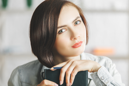 Close up portrait of attractive young woman with book, diary or notepad on blurry office background. Secretary concept の写真素材