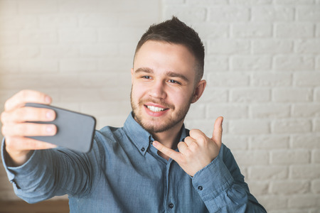 Portrait of attractive young man with call me gesture, taking selfie with smartphone in blurry office. Success and hipster concept の写真素材