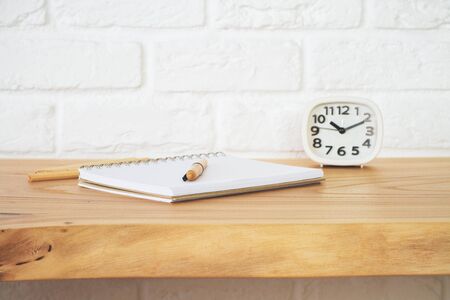 Close up of wooden office desktop with spiral notepad, pen, pencils and clock on white brick wall background. Supplies concept の写真素材