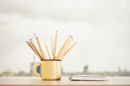 Close up of iron mug with pencils and spiral notepad on blurry city view background. Supplies, stationery items and tools concept の写真素材