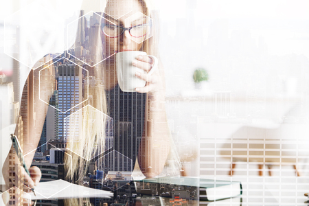 Portrait of pretty caucasian female drinking coffee and doing paperwork at abstract office workplace with city view and hexagonal pattern. Lifestyle and communication concept. Double exposure の写真素材