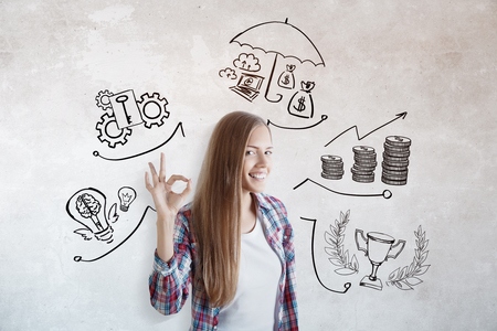 Portrait of attractive smiling european woman showing OK gesture on concrete wall background with creative business sketch. Success and investment conceptの写真素材