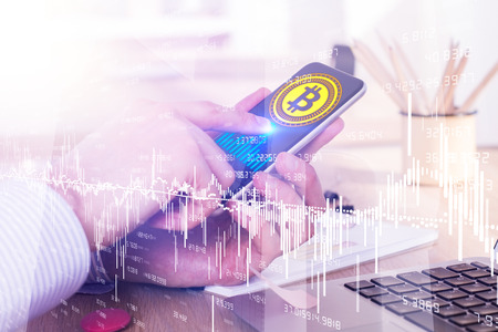 Side view of businessman hands holding and using smartphone with creative bitcoin hologram at desk...