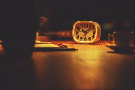 Night time workplace with clock, coffee cup, plant and other items illuminated by table lamp on blurry backgroundの写真素材