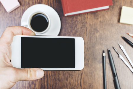 Hand holding empty smartphone over wooden desktop with supplies and coffee cup. Mock up の写真素材
