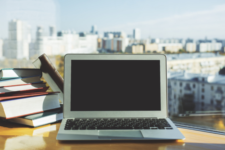 Front view of empty laptop placed on wooden desktop with books and blurry city view with sunlight. Mock up の写真素材