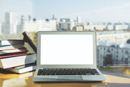 Front view of empty white laptop placed on wooden desktop with books and blurry city view with sunlight. Mock up の写真素材