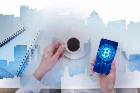 Top view of female hands using smartphone with bitcoin and holding coffee cup at abstract white desk with notepad and city skyline. E-commerce and finance concept. Double exposure の写真素材