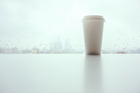Take out coffee cup placed on white windowsill with reflection. Window with rainy blurry morning city view in the backgrouns. Autumn or fall mood concept. Mock up の写真素材
