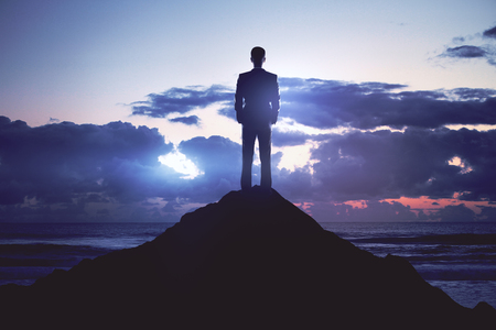 success concept with businessman standing on top of the rock and looking at cloudy skyの写真素材