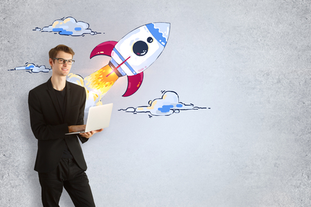 Businessman with laptop and creative launching rocket sketch on concrete wall background. Startup and technology conceptの写真素材