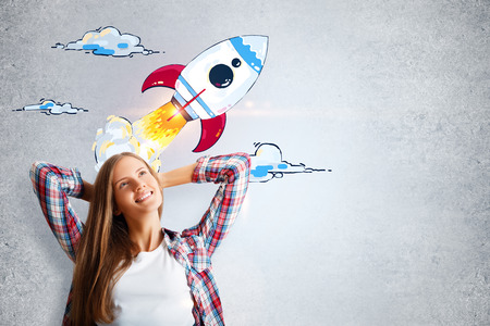 Attractive cheerful young woman daydreaming with creative launching rocket sketch on concrete wall background. Startup and success conceptの写真素材