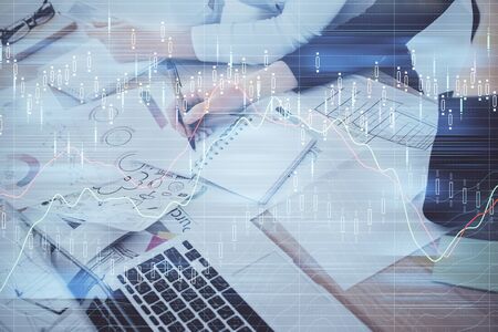 A woman hands writing information about stock market in notepad. Forex chart holograms in front....