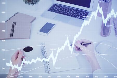 Hands writing information about stock market in notepad. Forex chart holograms in front. Concept of...