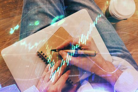 Double exposure of hands writing in notepad with stock market chart. Concept of research and...