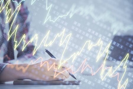 A woman hands writing information about stock market in notepad. Forex chart holograms in front....
