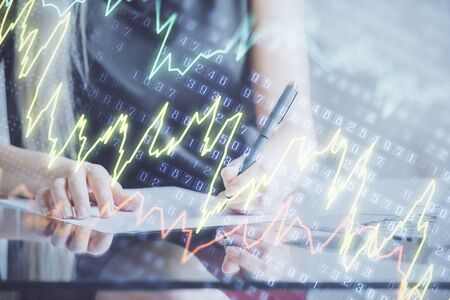 A woman hands writing information about stock market in notepad. Forex chart holograms in front. Concept of research.の写真素材