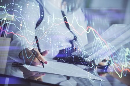 A woman hands writing information about stock market in notepad. Forex chart holograms in front....