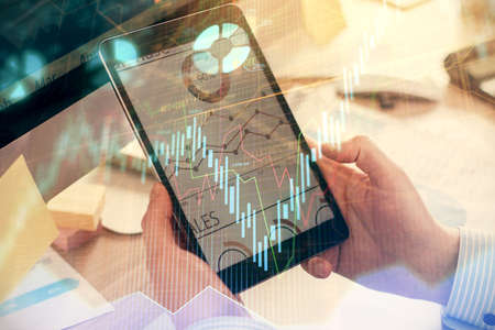 Double exposure of man's hands holding and using a phone and financial graph drawing. Analysis...