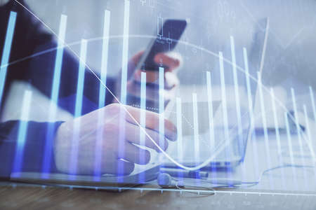 Double exposure of man's hands holding and using a phone and financial graph drawing. Analysis...