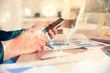 Double exposure of man's hands holding and using a phone and financial graph drawing. Analysis...