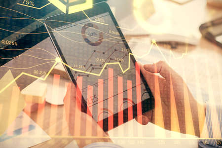 Double exposure of man's hands holding and using a phone and financial graph drawing. Analysis...