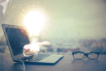 Computer on desktop in office with bulb icon hologram. Multi exposure. Concept of idea.