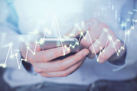 Double exposure of mans hands holding and using a digital device and forex graph drawing. Financial...