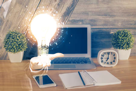Computer on desktop in office with bulb icon hologram. Multi exposure. Concept of idea.
