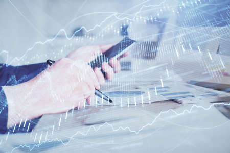 Double exposure of mans hands holding and using a phone and financial graph drawing. Analysis...