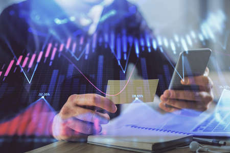 Double exposure of mans hands holding and using a phone and financial graph drawing. Analysis...