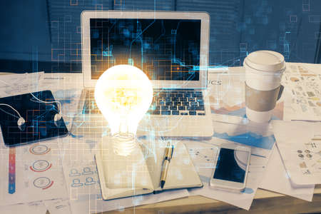 Computer on desktop in office with bulb icon hologram. Multi exposure. Concept of idea.