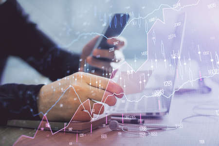 Double exposure of mans hands holding and using a phone and financial graph drawing. Analysis...