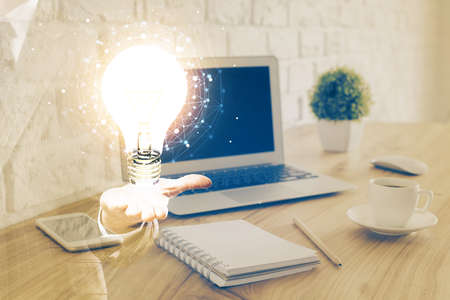 Computer on desktop in office with bulb icon hologram. Multi exposure. Concept of idea.