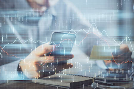 Double exposure of mans hands holding and using a phone and financial graph drawing. Analysis...