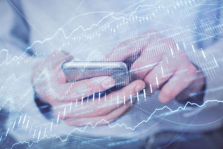 Double exposure of mans hands holding and using a digital device and forex graph drawing. Financial...