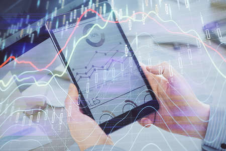 Double exposure of mans hands holding and using a phone and financial graph drawing. Analysis...