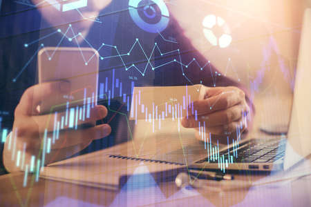 Double exposure of man's hands holding and using a phone and financial graph drawing. Analysis...