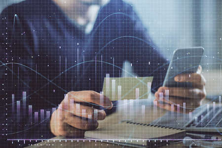 Double exposure of man's hands holding and using a phone and financial graph drawing. Analysis...