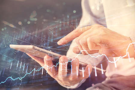 Double exposure of man's hands holding and using a digital device and forex graph drawing. Financial...